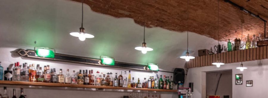Grün leuchtende Bankierslampen über der Flaschenwand, darüber das rohe Ziegelgewölbe.