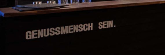 Auf die dunkle Holzfront der Theke ist in Schablonenschrift «Genussmensch sein.» gemalt.